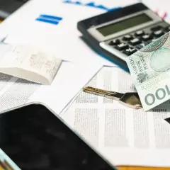 Dłoń trzyma banknot 100 zł nad dokumentami, kluczem i telefonem. To potwierdzenie odbioru gotówki.