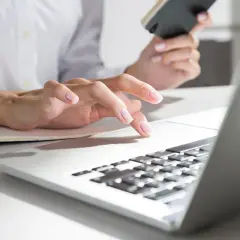 Dłoń kobiety pisząca na laptopie, trzymająca telefon.