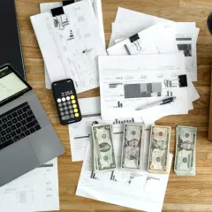 Laptop, telefon z kalkulatorem, dokumenty, pieniądze i lampka na biurku.