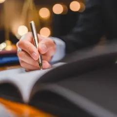 Dłoń pisze piórem w otwartym notatniku. W tle widać wagę sprawiedliwości i rozmyte światła, sugerujące formalne dokumenty, jak promesa zatrudnienia wzór.