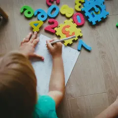 Dziecko pisze cyfry na kartce, otoczone piankowymi liczbami i sorterem.