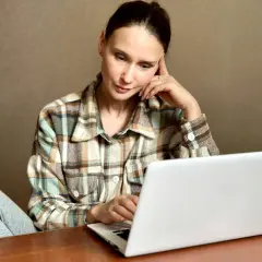 Kobieta w kratę siedzi przy laptopie, zamyślona, z ręką na skroni.