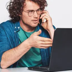 Mężczyzna w okularach rozmawia przez telefon, gestykulując w stronę laptopa. Szuka uczciwych firm oddłużeniowych, by rozwiązać swoje problemy finansowe.