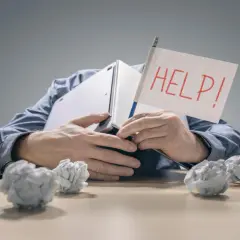 Zestresowany mężczyzna pod laptopem z flagą "HELP!" zastanawia się, ile wzrosną raty kredytów. Wokół niego papierowe kule.