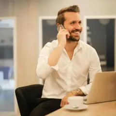 Uśmiechnięty mężczyzna w białej koszuli rozmawia przez telefon, siedząc przy biurku z laptopem. Może właśnie dowiaduje się, które banki dzwonią do pracodawcy.