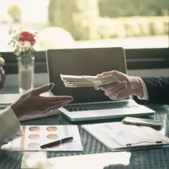 Dłoń przekazuje plik banknotów, co stanowi potwierdzenie przyjęcia gotówki. Na stole leżą dokumenty, laptop i telefon.