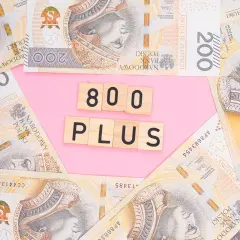 Polskie banknoty 200 zł i 100 zł ułożone na różowym tle, z drewnianymi klockami z napisem "800 PLUS".