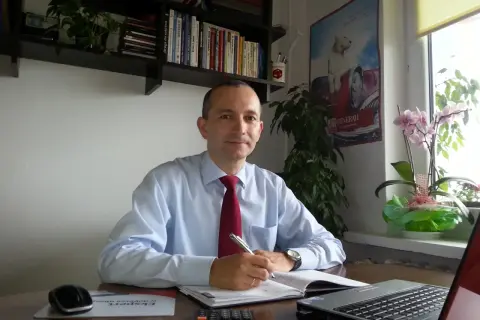 Mężczyzna w jasnej koszuli i czerwonym krawacie siedzi przy biurku, pisząc w notatniku.