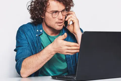 Mężczyzna w okularach rozmawia przez telefon, gestykulując w stronę laptopa. Szuka uczciwych firm oddłużeniowych, by rozwiązać swoje problemy finansowe.