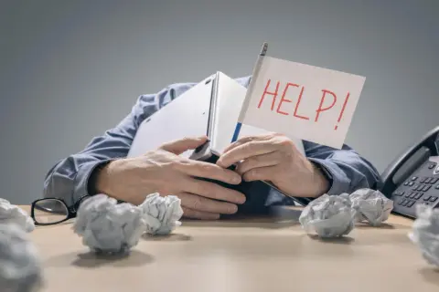 Zestresowany mężczyzna pod laptopem z flagą "HELP!" zastanawia się, ile wzrosną raty kredytów. Wokół niego papierowe kule.