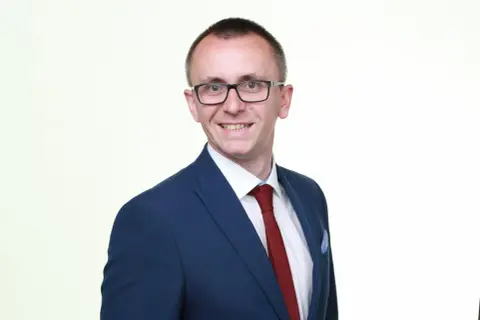 Mężczyzna w okularach, granatowej marynarce i czerwonym krawacie, uśmiecha się do aparatu.