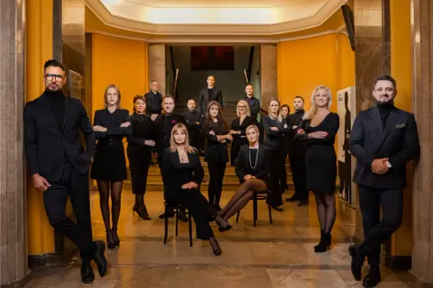 Grupa profesjonalistów w czarnych strojach pozuje na tle żółtej ściany i schodów.