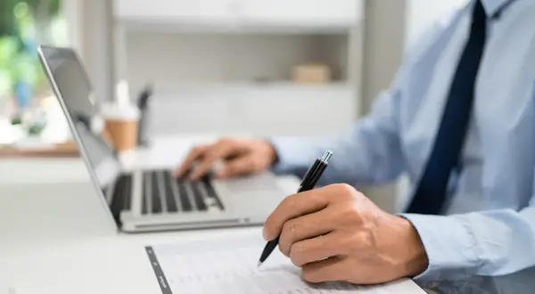 Mężczyzna w biurze pracuje przy laptopie i wypełnia dokumenty. Dowiaduje się, jak złożyć wniosek o wakacje kredytowe.