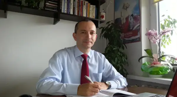 Mężczyzna w jasnej koszuli i czerwonym krawacie siedzi przy biurku, pisząc w notatniku.