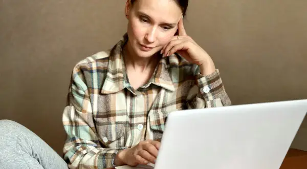 Kobieta w kratę siedzi przy laptopie, zamyślona, z ręką na skroni.