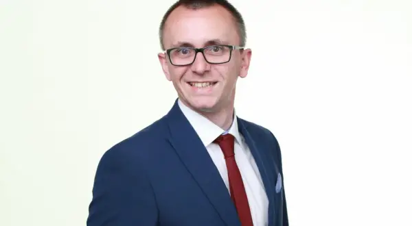 Mężczyzna w okularach, granatowej marynarce i czerwonym krawacie, uśmiecha się do aparatu.
