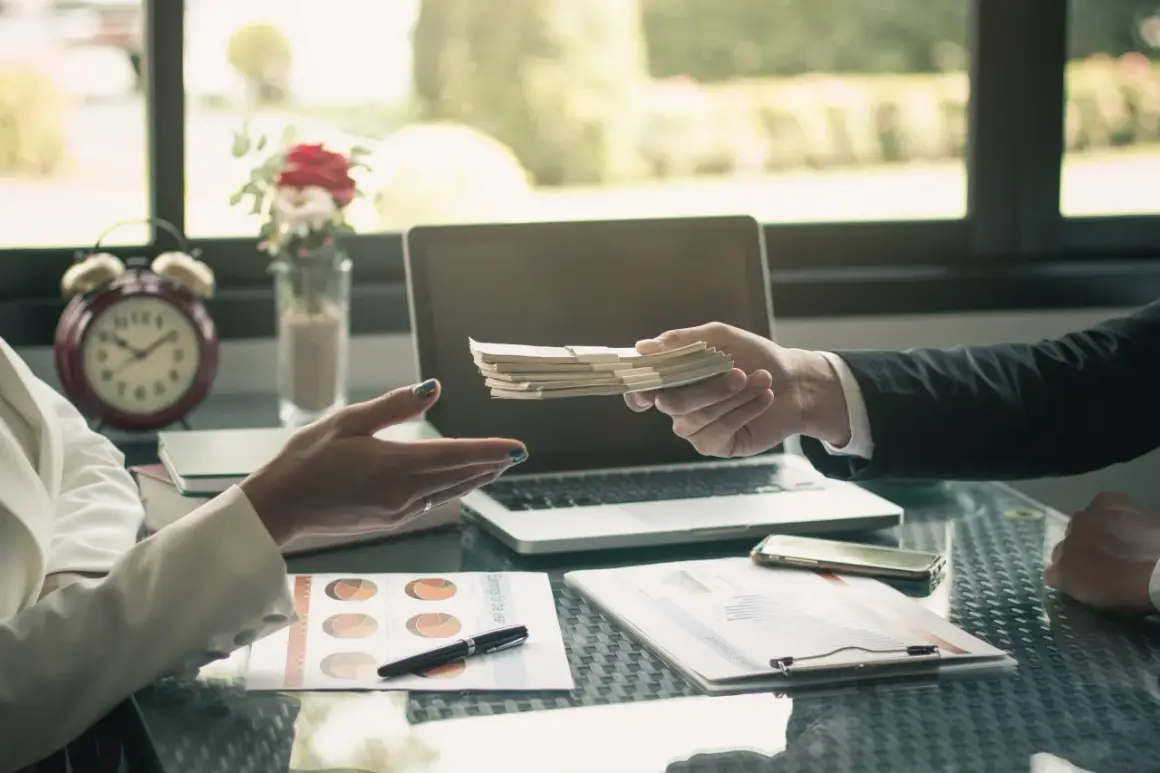 Dłoń przekazuje plik banknotów, co stanowi potwierdzenie przyjęcia gotówki. Na stole leżą dokumenty, laptop i telefon.