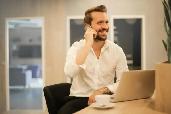 Uśmiechnięty mężczyzna w białej koszuli rozmawia przez telefon, siedząc przy biurku z laptopem. Może właśnie dowiaduje się, które banki dzwonią do pracodawcy.